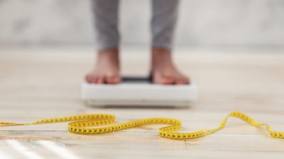 A Black woman stands on a scale out of focus. A measuring tape is in focus.