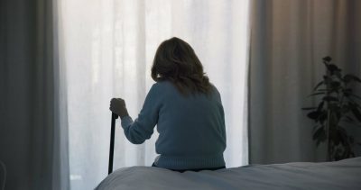 An image of an elderly woman sits on the edge of a bed and looks out a window.