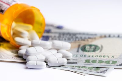 close up of a prescription bottle with white tablets falling out on top of a group of hundred dollar bills
